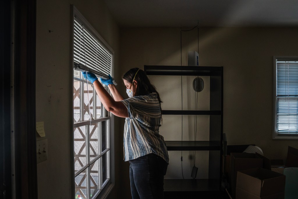 A person wearing a face mask and blue gloves stands inside a sparsely furnished apartment, lifting the blinds to look out a window. Empty shelves and moving boxes are visible in the dimly lit room behind them.