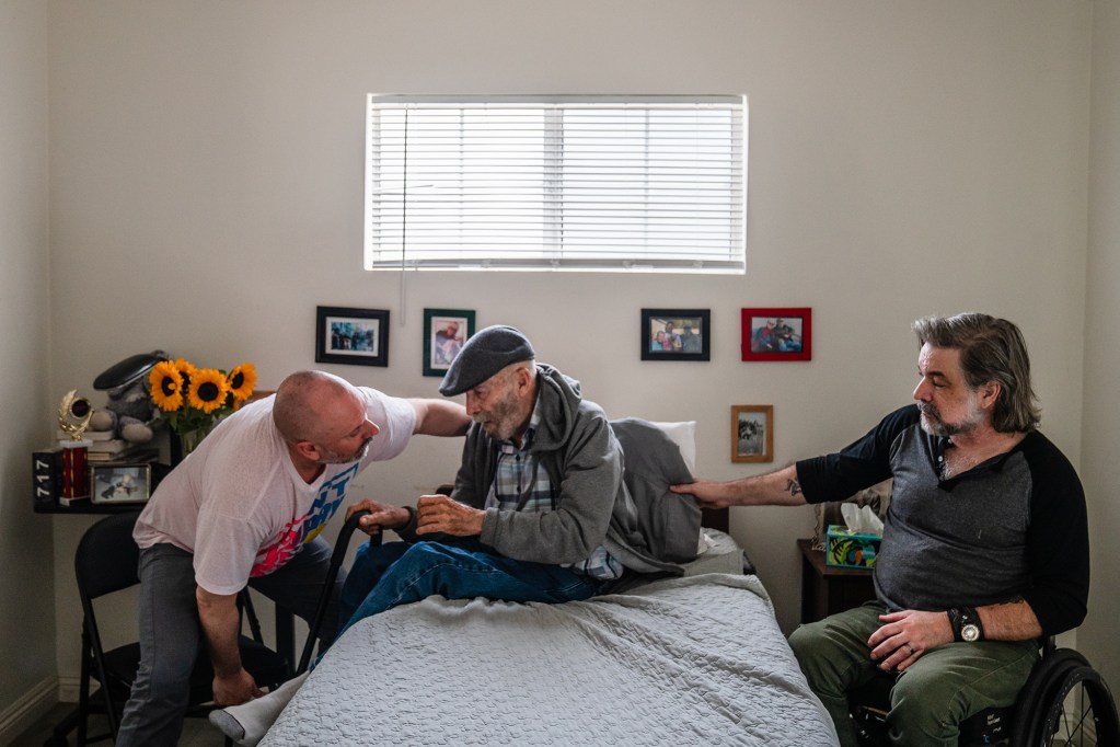 Two people help and elderly person get up from their bed inside a room with framed photos hung up on the wall next to a window.