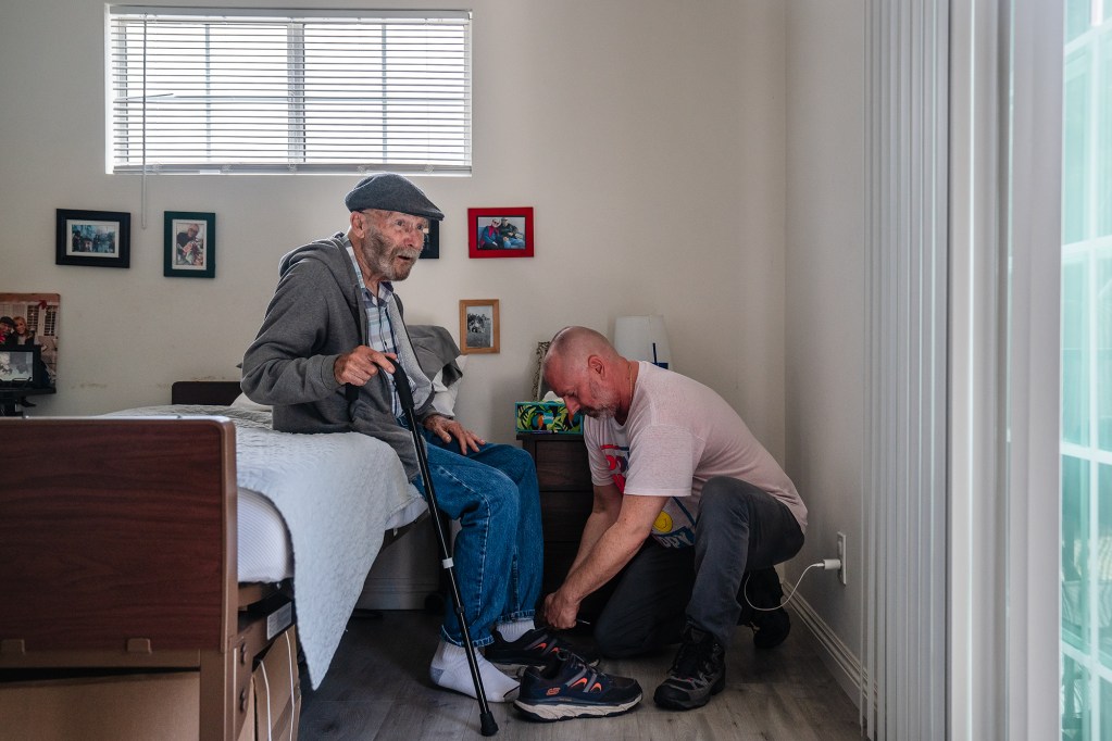 A person wearing a pink shirt and gray pants, ties an elderly person's shoe as they sit on the edge of a bed inside a room with framed photos hung up on the wall next to a window.