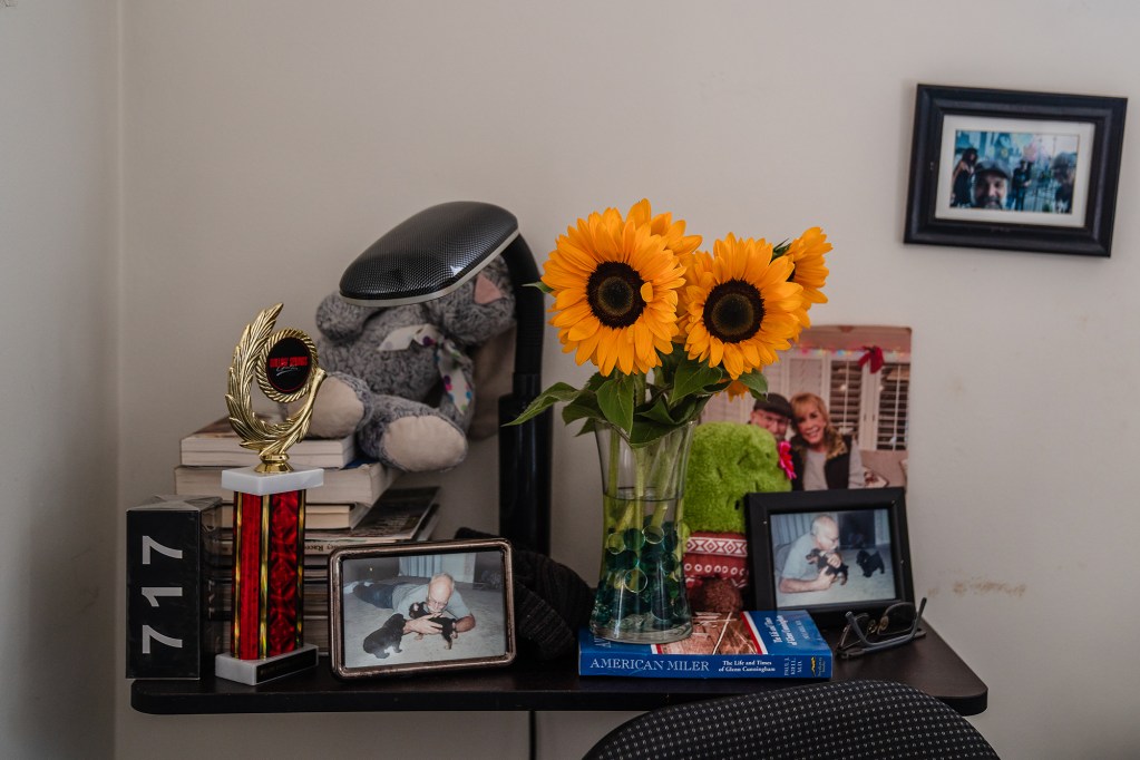 A nightstand with framed family photos, books, trophies and a vase with sunflowers.