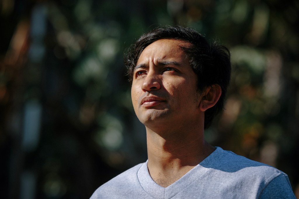 A person in a gray T-shirt looks upward while standing outdoors in bright sunlight. Their face is sharply lit against a dark, softly blurred background of trees and foliage.