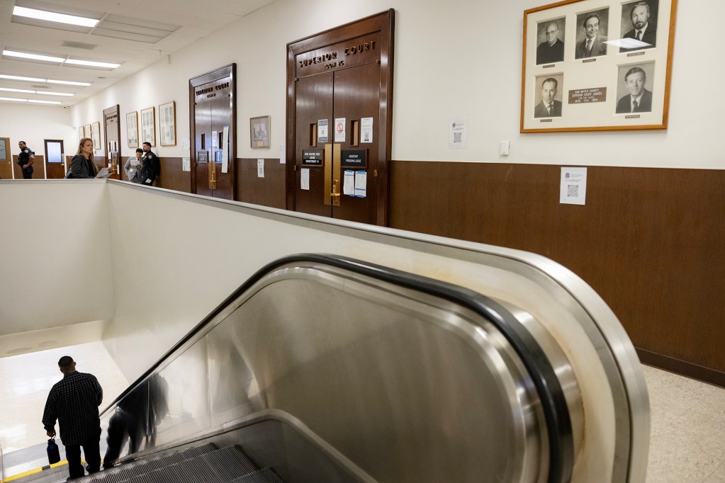 A person carrying a water bottle and other belongings descends an escalator inside a courthouse, passing several Superior Court courtroom entrances. Police officers and other people stand farther down the hallway.