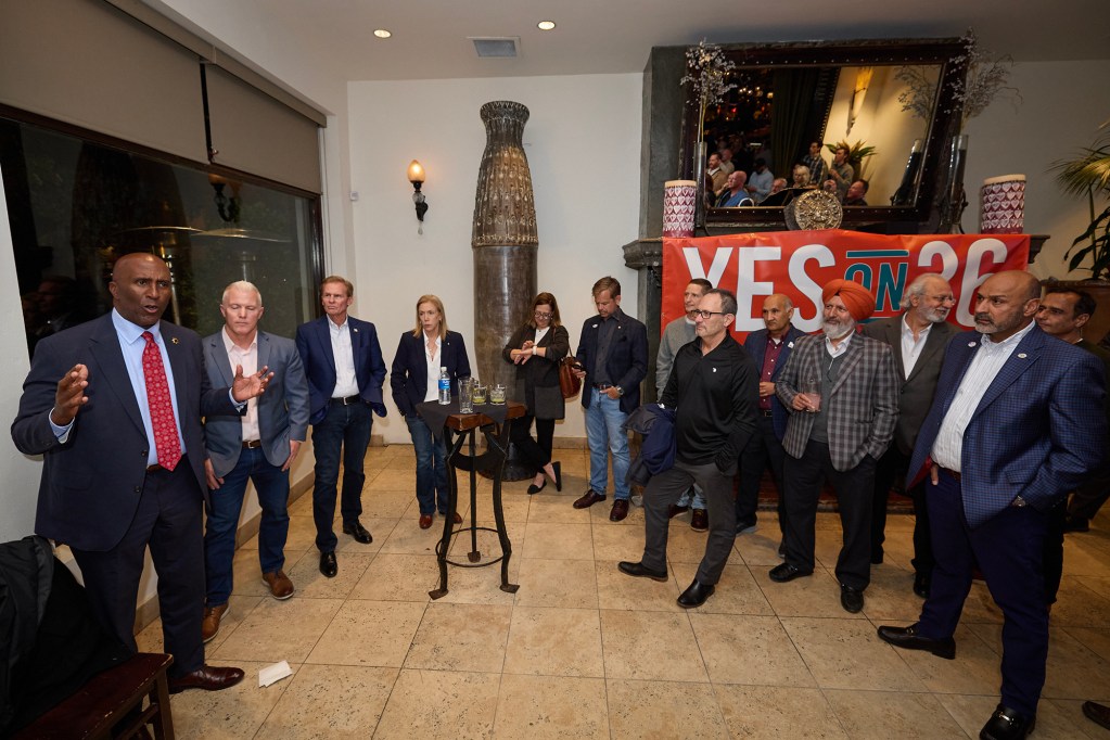 A group of people gathers indoors in front of a large “YES ON 36” campaign banner as a person in a suit speaks to the crowd. Attendees stand around the room listening, while additional people are reflected in a large mirror behind the group.