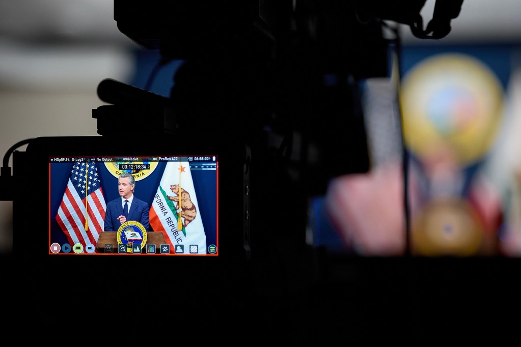 Gov. Gavin Newsom speaks at a podium between American and California state flags, seen through the monitor of a television camera during a press conference. The camera equipment is silhouetted in the foreground.