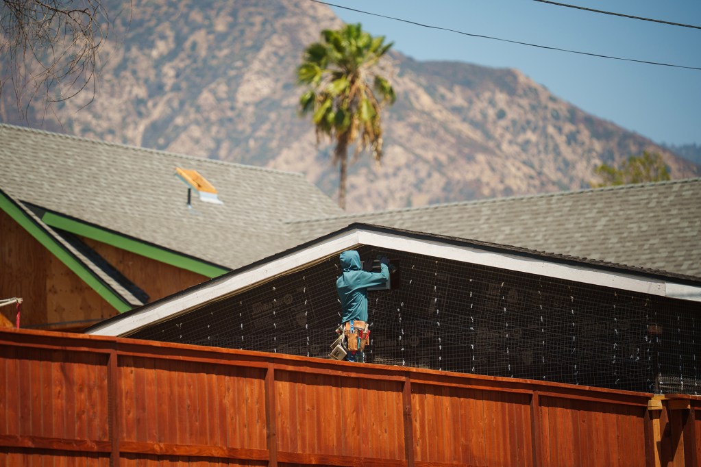 A construction worker in a teal hoodie works beneath the roofline of a house behind a reddish-brown wooden fence, with a palm tree and rugged mountains in the background.