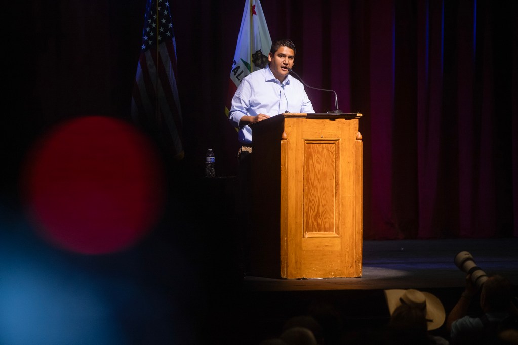 Randy Villegas speaks from a wooden podium onstage, with American and California flags behind him and audience members in the foreground.