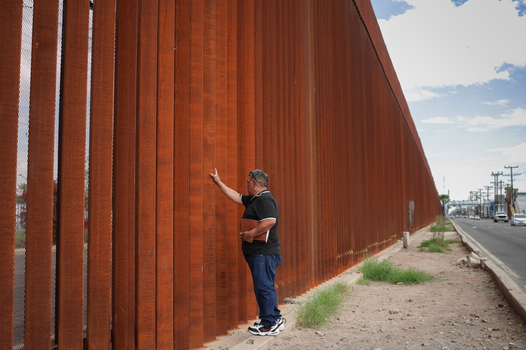 Luis Arreguin touches a tall, rust-colored steel border wall while holding a Bible under his arm. The wall stretches into the distance alongside a road.
