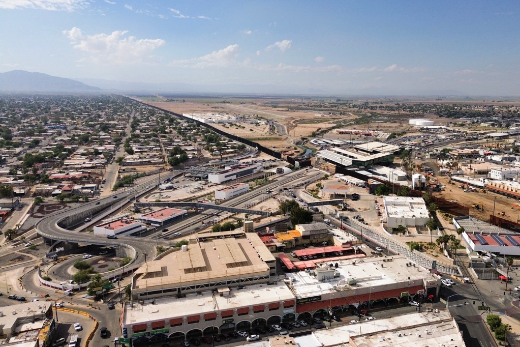 An aerial view of the U.S.-Mexico border crossing between Calexico and Mexicali, with roads, inspection facilities and neighborhoods surrounding the border wall.